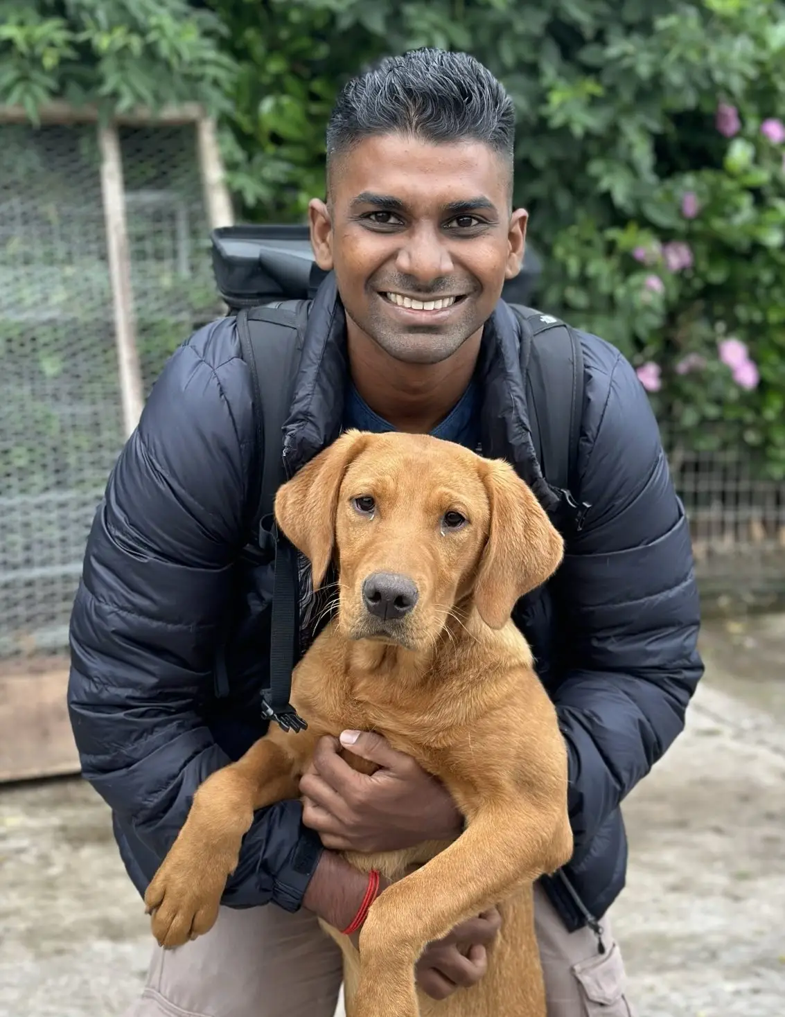 A smiling Singaporean dog trainer in a black puffer jacket and cargo pants holds a golden-brown Labrador Retriever dog